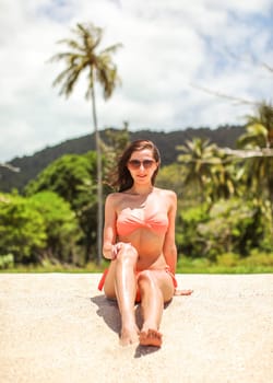 Young sporty woman in orange bikini and sunglasses sits on fine beach sand, wind in her hair, palm tree and jungle behind her.