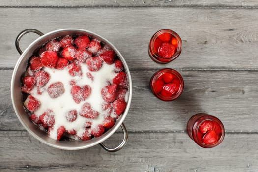 Tabletop view, steel pot with strawberries crystal sugar on top, three bottles with pickled fruit next to it. Homemade strawberry compote preparation.