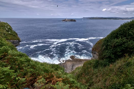 A lighthouse on the Cantabrian Sea coast, and coast line, Calm waters, waves on the shore, vegetation, cliffs, sailboats, seagulls