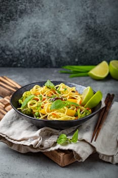 Asian vegetarian noodles with vegetables and lime in black rustic ceramic bowl, wooden chopsticks on cutting board angle view on stone background. Cooking noodles concept