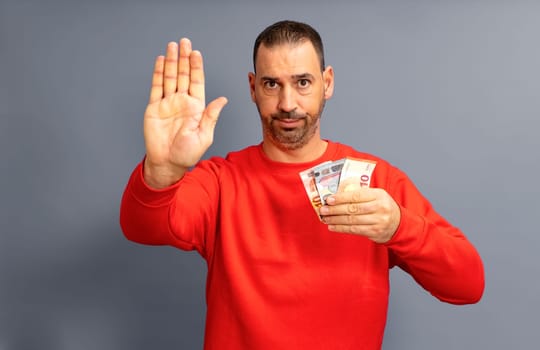 Hispanic man in his 40s holding a euro bill isolated on gray background standing with outstretched hand showing stop sign, preventing you