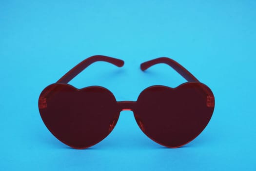 Dark red female sunglasses front view. ..Elegant red plastic glasses in the shape of a heart on a blue paper background close-up