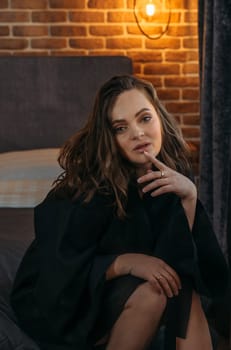 Portrait of a beautiful woman in a black bathrobe, who is sitting on the bed, looking at the camera.