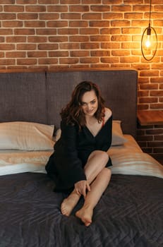 Portrait of a beautiful woman in a black bathrobe, who is sitting on the bed with her legs tucked under her, looking at the camera.