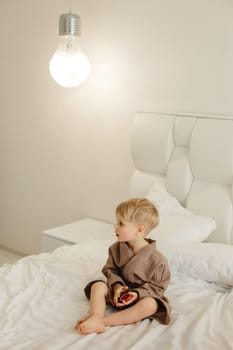 Portrait of a boy in a brown bathrobe, who is sitting on a white bed with candied oranges in his hands.