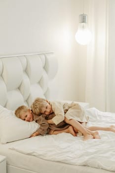 The brothers lie in dressing gowns in a white bed, play, hug, smile. Family concept.