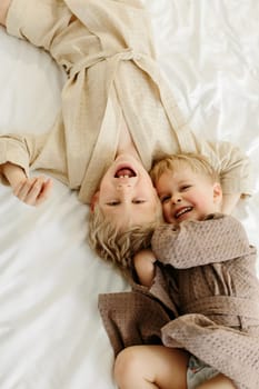 The brothers lie in dressing gowns in a white bed, play, hug, smile. Family concept. View from above.