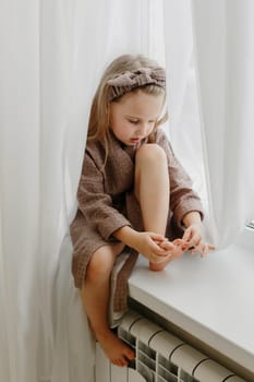 Portrait of a girl in a brown dressing gown, who sits on the windowsill and examines her toes.