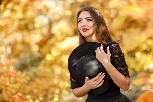 beautiful woman in black dress and hat on a background of yellow autumn leaves