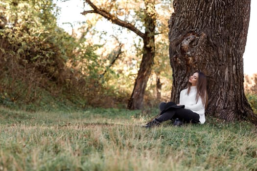 beautiful woman sitting under big tree and daydreaming on sunny autumn day.