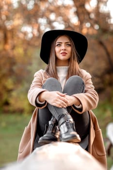 beautiful fashion woman in beige coat and black hat sitting on tree on sunny autumn day.