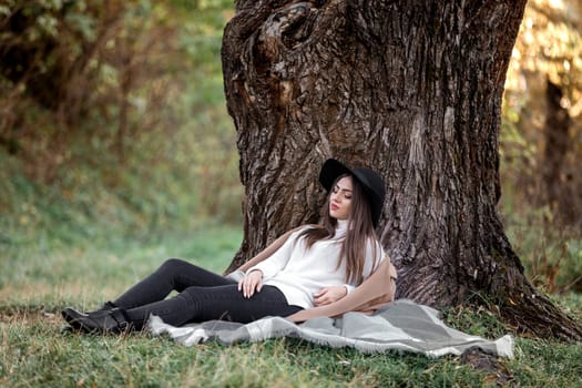 beautiful woman in a beige coat and black hat sitting under big tree on sunny autumn day.