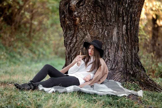 beautiful woman in a beige coat and black hat sitting under big tree on sunny autumn day.