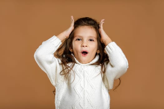 happy surprised beautiful curly little child girl in white sweater looking to camera on beige background. Human emotions and facial expression