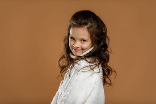 Cute shy little child girl in white sweater looking to camera on beige background. Human emotions