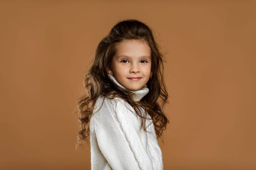 Cute smiling little child girl in white sweater looking to camera on beige background. Human emotions and facial expression