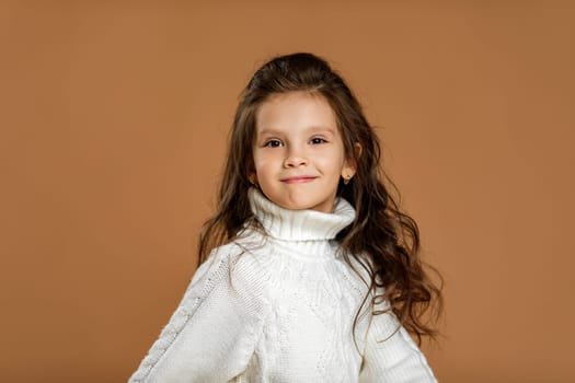 Cute smiling little child girl in white sweater looking to camera on beige background. Human emotions and facial expression