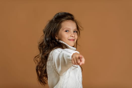 Cute smiling curly little girl in white sweater pointing camera on beige background. child pointing at you