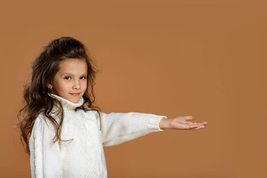 Cute smiling curly little girl in white sweater pointing at something on beige background. place your product here