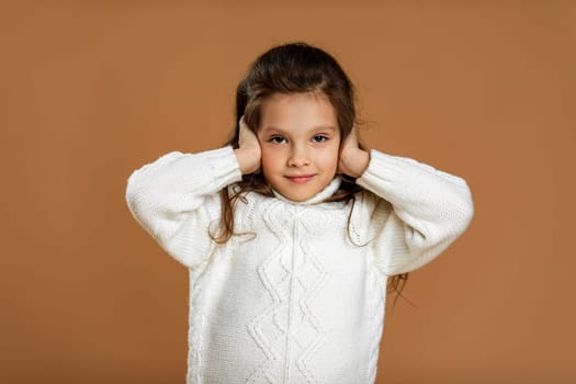 Cute curly little girl in white sweater is closing her ears with her hand on beige background.