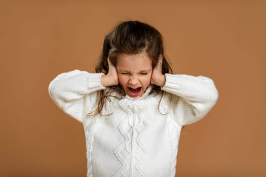 Cute curly little girl in white sweater is closing her ears with her hand and screaming on beige background.