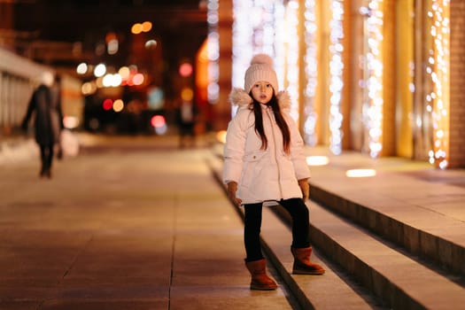 Cute happy little girl walks on the streets of a big city in the evening in winter. Christmas holidays.