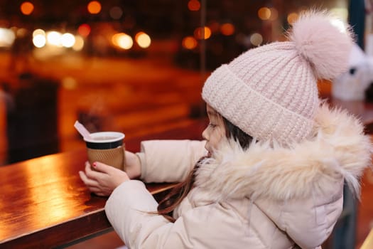 Cute happy little girl drinks hot tea in a street cafe in the evening in winter. child is cold. Christmas holidays