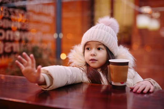 Cute happy little girl drinks hot tea in a street cafe in the evening in winter. child is cold. Christmas holidays