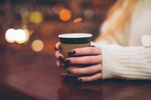 female hands holding a paper cup with hot coffee in a cafe on a cold winter evening