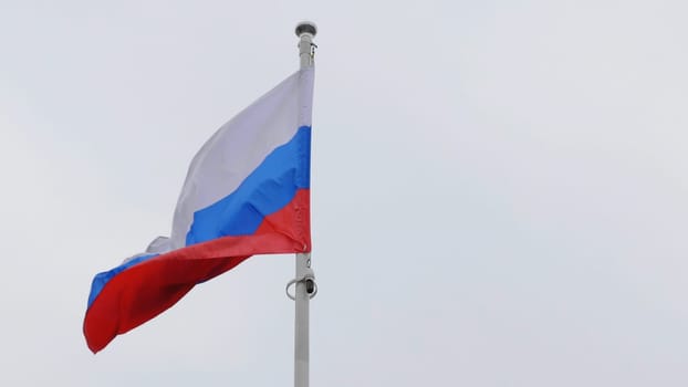Russian flag on the flagpole waving in the wind against a blue sky with clouds.