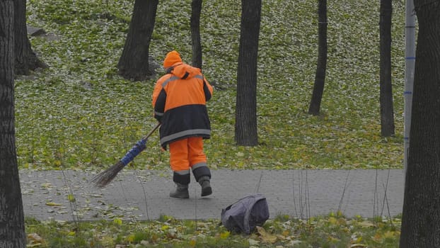 Worker sweeps autumn leaves with a broom from the sidewalk.