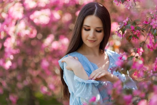 Pretty girl with long hair is standing near a pink blooming apple tree, in the summer in the park. Close up