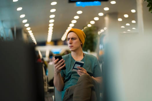 A young man sits at the airport and checks in for the next flight via an app on his phone, waits for the plane and works while traveling.