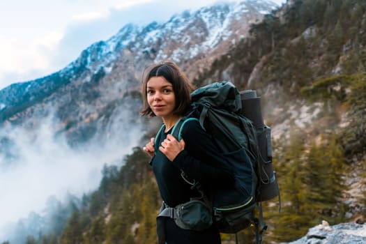 Young smiley girl with backpack and climbing equipment walks on the mountain trail in the national park. The hiker explores wildlife, woodland, beautiful foggy forest and mountains.