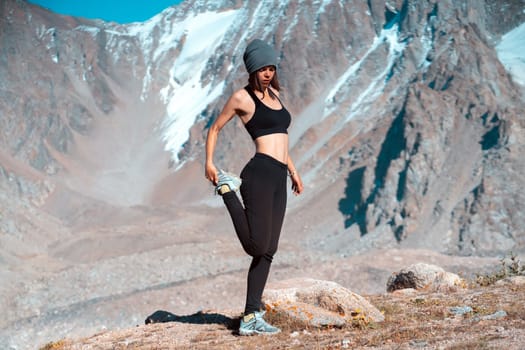 A young sports girl is warming up on a background of snow-capped mountains on a sunny day, a woman does exercises, trains and meditates in a picturesque mountainous area.