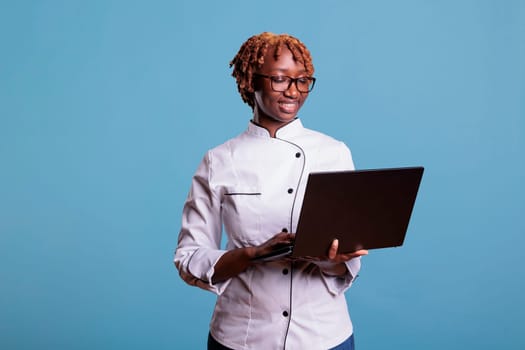 African american woman chef using a computer, searching, browsing recipes on Internet. Studio portrait of female afro hair cooker, using laptop to work video call, conference.