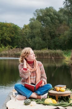 a beautiful red-haired girl reads a book on the embankment against the backdrop of autumn yellowing forests, fashion sweater. High quality photo