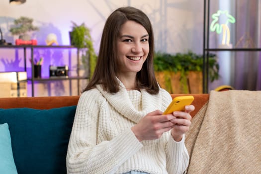 Portrait of woman sits on sofa uses mobile phone smiling at home living room apartment. Girl texting share messages content stories on smartphone social media applications online watching relax movie