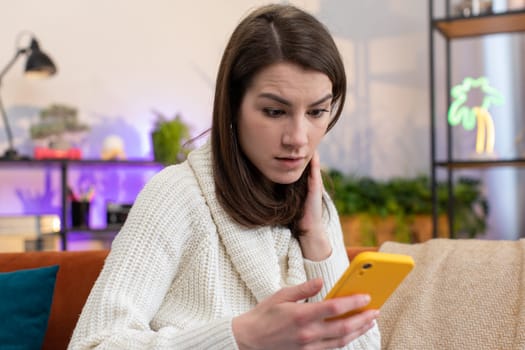 Sad displeased woman use smartphone typing browsing, loses becoming surprised sudden lottery results, bad news, fortune loss, fail, deadline, virus. Portrait of unlucky girl at home room sits on couch