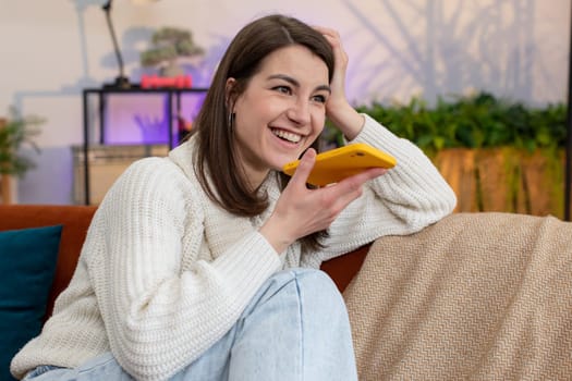 Portrait of happy excited young woman enjoying mobile smartphone loudspeaker talking good news, laughing at home room apartment. Girl making phone call conversation with friends sitting on couch