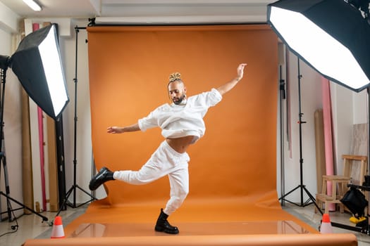 Full length african-american gay man with bright make up jumping wearing white dancing on orange studio background. Brazilian male posing in photo studio. Bearded afro-american gay man dancer