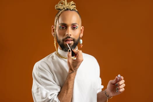 African american gay applying make up isolated on orange background paint lips with lipstick isolated brown background. Fashion lgbtq concept.