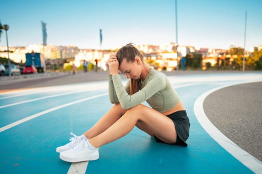 Tired happy woman runner taking rest after run sitting on the running track and smile. Summer evening time. Young athletic female jogger resting after training. Work hard rest harder. Copy space