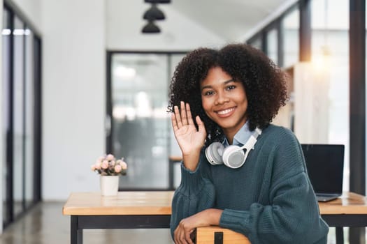 Happy African American student in wireless headphones waving hand hello with toothy smile, laughing at camera, making video call at home. Black woman talking online head shot portrait screen.