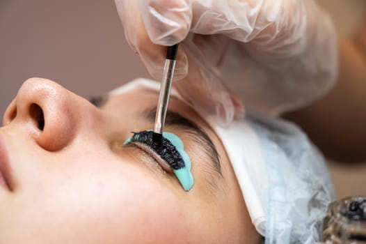 Close-up portrait of a woman on eyelash lamination procedure. The master applies tint to the eyelashes