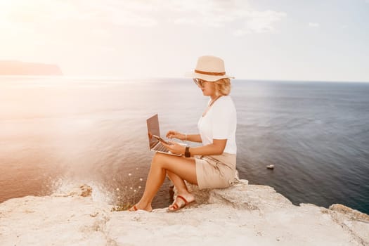 Digital nomad, Business woman working on laptop by the sea. Pretty lady typing on computer by the sea at sunset, makes a business transaction online from a distance. Freelance remote work on vacation