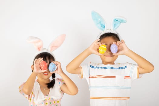 Happy Easter Day. Two smile Asian little girl and boy wearing easter bunny ears holding colorful eggs closes eyes with testicles isolated on white background with copy space, Happy child in holiday
