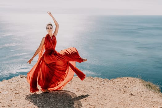 Side view a Young beautiful sensual woman in a red long dress posing on a rock high above the sea during sunrise. Girl on the nature on blue sky background. Fashion photo.