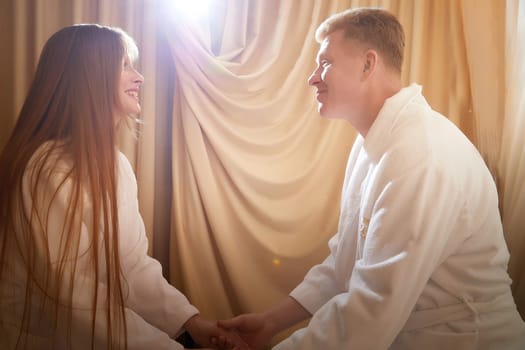 An adult couple of newlyweds in in white robes in a hotel room after the wedding. A guy and a girl, a man and a woman in beautiful room. The concept of hugs, love and care