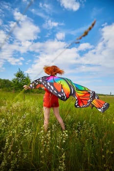 Adult girl with red hair and butterfly wings having fun and joy in meadow or field with grass, flowers on sunny summer day